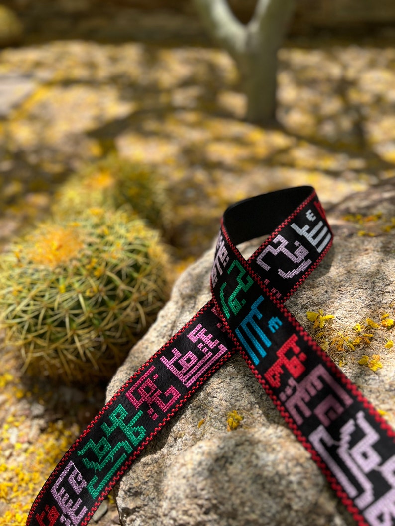 Peut inclure: Un bracelet noir tiss&eacute; avec du texte arabe brod&eacute; en couleur. Le bracelet est pos&eacute; sur un rocher avec un cactus en arri&egrave;re-plan.