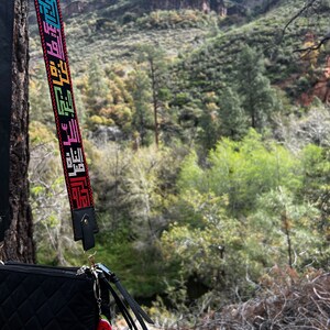 Peut inclure: Un sac &agrave; bandouli&egrave;re noir matelass&eacute; avec une sangle color&eacute;e orn&eacute;e d'un motif textuel r&eacute;p&eacute;titif. Le sac a un charme de fraise attach&eacute; au tirette de la fermeture &eacute;clair. Le sac est suspendu &agrave; une branche d'arbre devant un paysage montagneux.