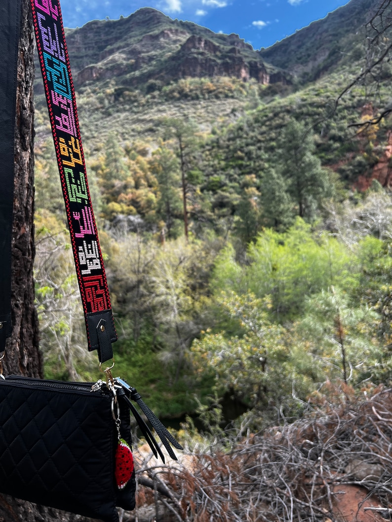 Peut inclure: Un sac &agrave; bandouli&egrave;re noir matelass&eacute; avec une sangle color&eacute;e orn&eacute;e d'un motif textuel r&eacute;p&eacute;titif. Le sac a un charme de fraise attach&eacute; au tirette de la fermeture &eacute;clair. Le sac est suspendu &agrave; une branche d'arbre devant un paysage montagneux.