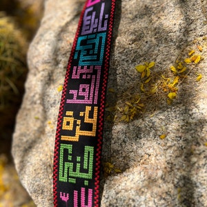 Peut inclure: Une sangle tiss&eacute;e noire avec des inscriptions arabes brod&eacute;es color&eacute;es. La sangle est faite d'un mat&eacute;riau &eacute;pais et durable et a une bordure rouge.