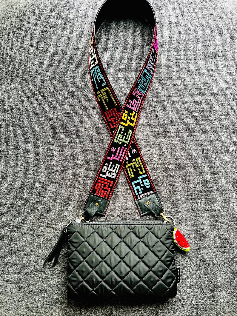 Peut inclure: Un sac &agrave; bandouli&egrave;re matelass&eacute; noir avec une bandouli&egrave;re noire et multicolore orn&eacute;e d'&eacute;criture arabe. Le sac a un charme past&egrave;que attach&eacute; &agrave; lui.