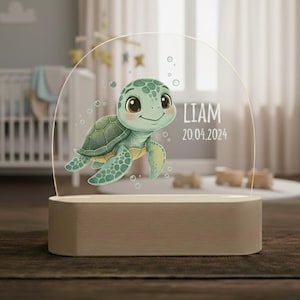 Könnte beinhalten: Dekorative Nachtlampe mit einem Cartoon-Design einer Meeresschildkröte. Die Schildkröte ist grün und gelb, mit Blasen um sie herum. Der Name "LIAM" und das Datum "20.04.2024" sind auf dem transparenten Acryl aufgedruckt. Der Sockel ist hellbeige.
