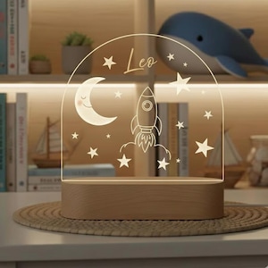 Könnte beinhalten: Dekoratives Nachtlicht mit Holzsockel und klarem Acrylbogen. Der Bogen zeigt eine Rakete, einen Mond, Sterne und den Namen "Leo". Ideal für ein Kinderzimmer.