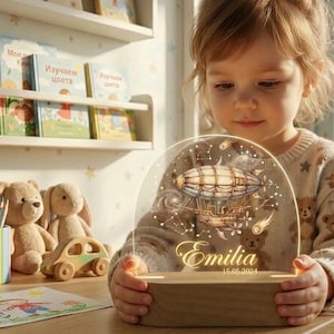 Könnte beinhalten: Ein Nachtlicht mit Holzsockel und einem klaren, gewölbten Oberteil mit einem Vintage-Luftschiff-Design, Sternbildern und dem Namen "Emilia" mit dem Datum 15.05.2024. Das Licht wird von einem Kind gehalten. Weitere Spielsachen und Bücher sind im Hintergrund.