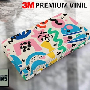 May include: A colorful case with an abstract design featuring shapes in pink, blue, yellow, and green. The case has the text "3M PREMIUM VINIL" and the logo "WUNDERBAR SKINS".