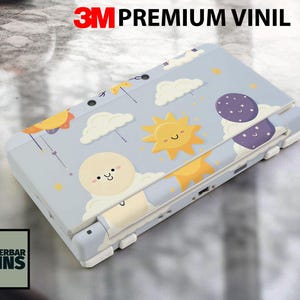 May include: A light blue Nintendo 3DS skin featuring a whimsical design of a smiling sun, clouds, and a smiling moon. The skin is labeled "3M Premium Vinil" and "Wunderbar Skins".