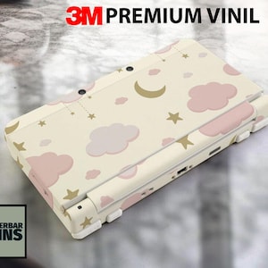 May include: A cream-colored handheld gaming console skin featuring a pattern of pink clouds, gold stars, and a crescent moon. The text "3M PREMIUM VINIL" is at the top. The brand "WUNDERBAR SKINS" is in the lower left corner.