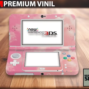 May include: Pink and white Nintendo 3DS console with a cloud design. The console is closed and the top screen displays the Nintendo 3DS logo. The console is on a wooden surface.