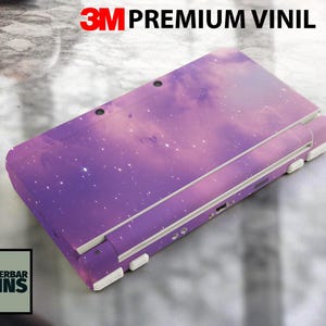 Puede incluir: Una funda para consola de juegos portátil con temática de galaxia púrpura y rosa. La funda presenta un diseño de nebulosa con estrellas blancas. El texto "3M PREMIUM VINIL" está en la parte superior. El logotipo de "WUNDERBAR SKINS" está en la parte inferior.