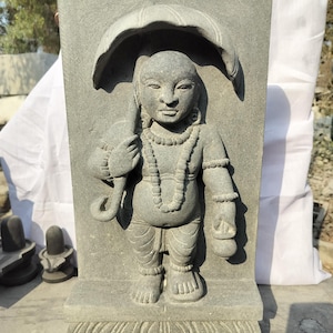 Puede incluir: Escultura de piedra gris de una figura de pie sosteniendo un gran paraguas. La figura está adornada con un collar y pulseras. La escultura está sobre una base rectangular con un diseño floral.