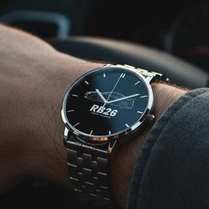 May include: A silver-tone wristwatch with a round black dial featuring a white line-art graphic of a car and the text RB26 TWIN TURBO. The watch has a metallic link bracelet and is worn on a wrist while holding a black steering wheel.