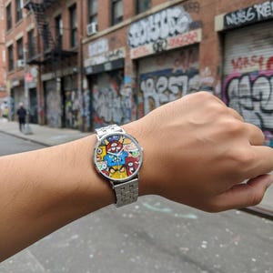 May include: A silver wristwatch with a colorful, cartoon-style face featuring various characters in blue, red, and yellow. The watch has a silver band and is worn on a wrist.