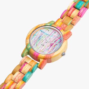 May include: A wooden wristwatch with a colorful, woven band. The watch face has a white background with a colorful scribble design and silver hands. The watch case and band are made of wood in various colors.