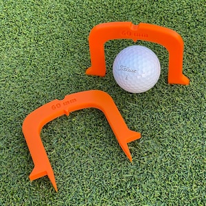 Puede incluir: Dos ayudas de entrenamiento de golf de plástico naranja sobre hierba verde. La ayuda más grande tiene 60 mm de ancho y la más pequeña 50 mm de ancho. Una bola de golf blanca descansa sobre la hierba entre las dos ayudas.