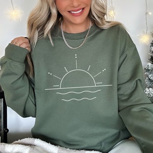 May include: A green sweatshirt featuring a white graphic of a sun setting over water. The graphic is a simple line drawing with a circle for the sun and wavy lines for the water.