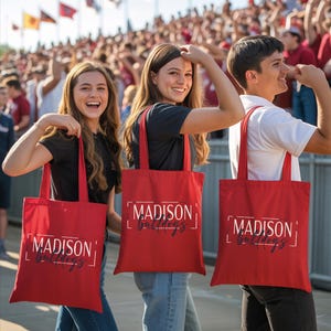 Three people holding red tote bags with the word "Madison" printed on them, standing in front of a crowd at an event.