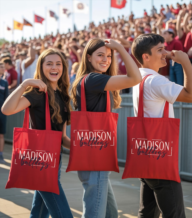 Three people holding red tote bags with the word "Madison" printed on them, standing in front of a crowd at an event.