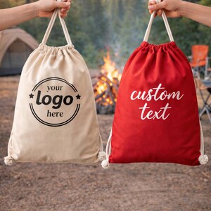 Two hands holding fabric bags with custom text printed on them, one beige and one red, in front of a campfire and pine trees.