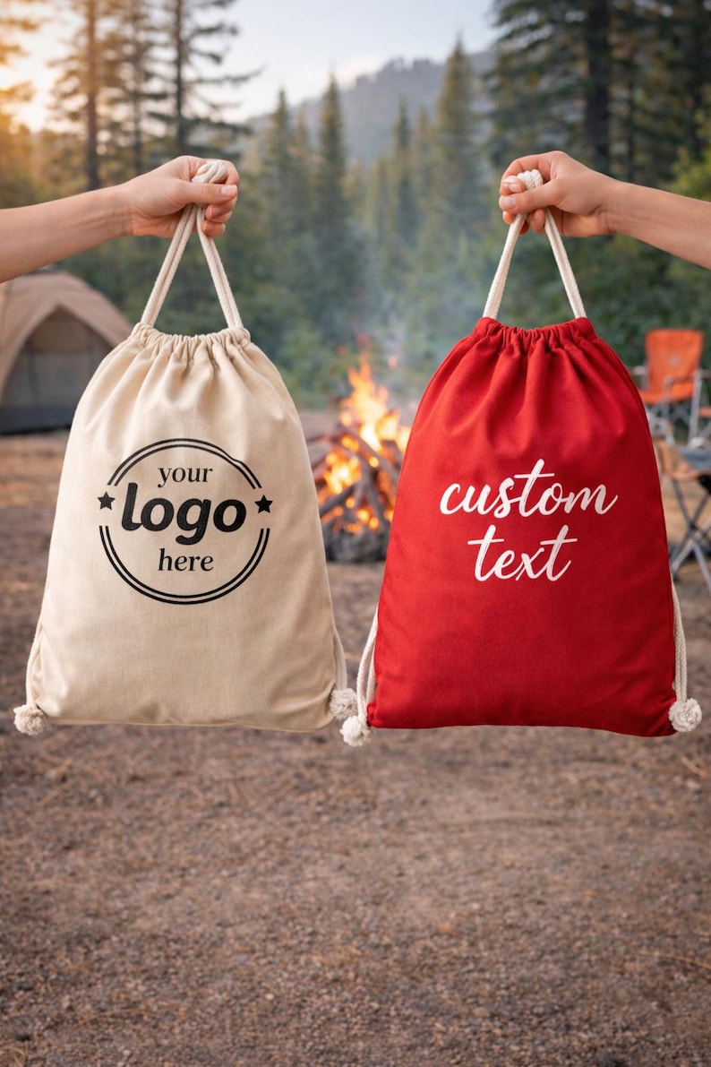 Two hands holding fabric bags with custom text printed on them, one beige and one red, in front of a campfire and pine trees.