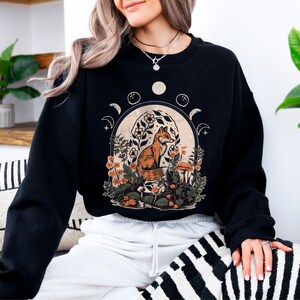 May include: Black sweatshirt with a graphic print of a fox sitting in a forest scene with flowers and mushrooms. The graphic also includes a crescent moon and stars.