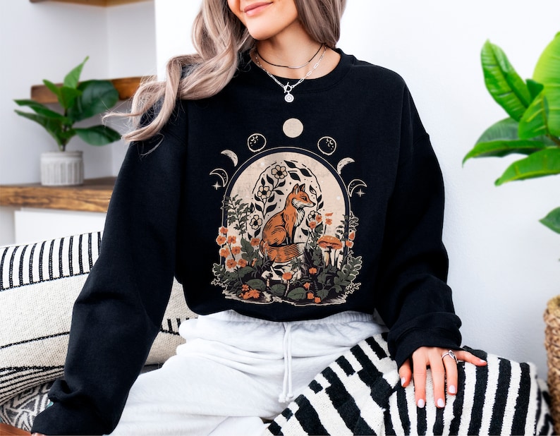 May include: Black sweatshirt with a graphic print of a fox sitting in a forest scene with flowers and mushrooms. The graphic also includes a crescent moon and stars.