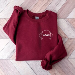 May include: Burgundy sweatshirt with a white outline of a dog's head and the name "Tucker" written inside.
