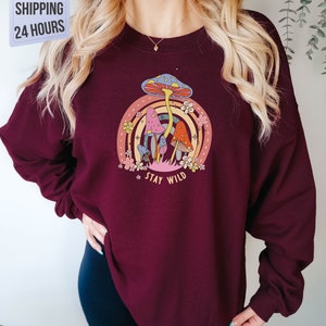 May include: A maroon sweatshirt with a graphic of colorful mushrooms and a rainbow. The text "Stay Wild" is printed below the mushrooms.