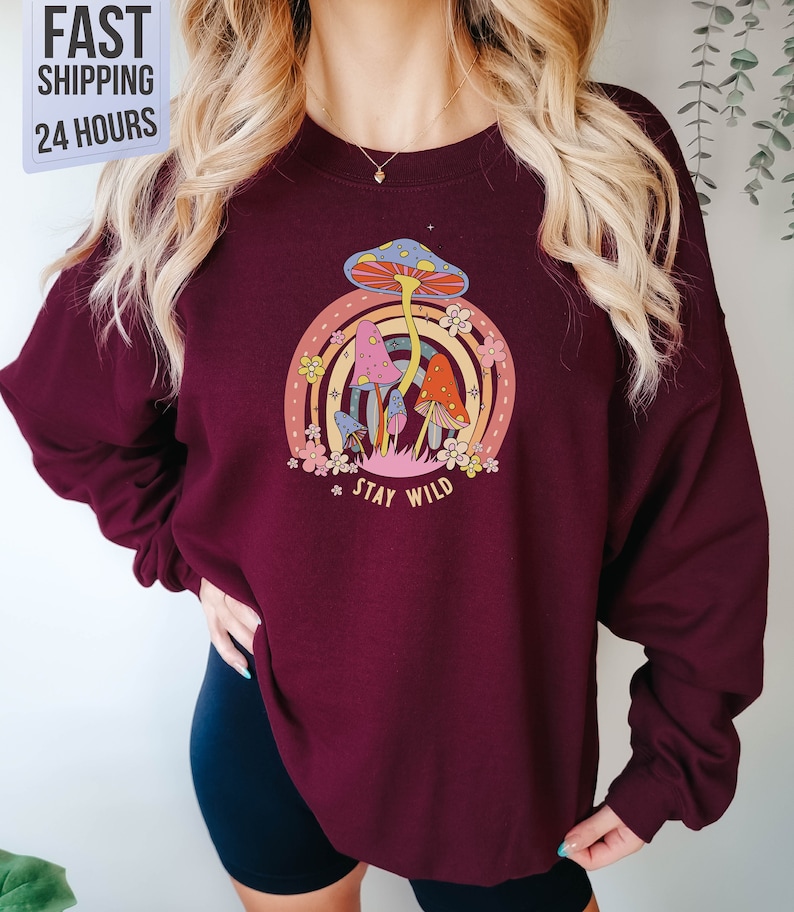 May include: A maroon sweatshirt with a graphic of colorful mushrooms and a rainbow. The text "Stay Wild" is printed below the mushrooms.