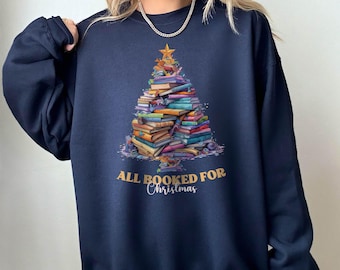 May include: A navy blue crewneck sweatshirt featuring a graphic of a Christmas tree constructed from a stack of colorful books. A gold star sits at the top of the book tree. The text "ALL BOOKED FOR Christmas" is printed in gold and white.