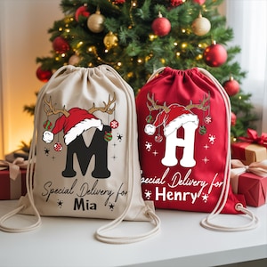 May include: Two drawstring Christmas gift bags. One is beige with a black "M" and the text "Special Delivery for Mia". The other is red with a white "H" and the text "Special Delivery for Henry". Both bags have a Santa hat and reindeer antlers design.