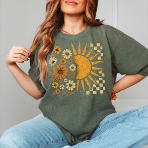 A woman wearing a green t-shirt with a sun and flower design, sitting with her hands on her hips.