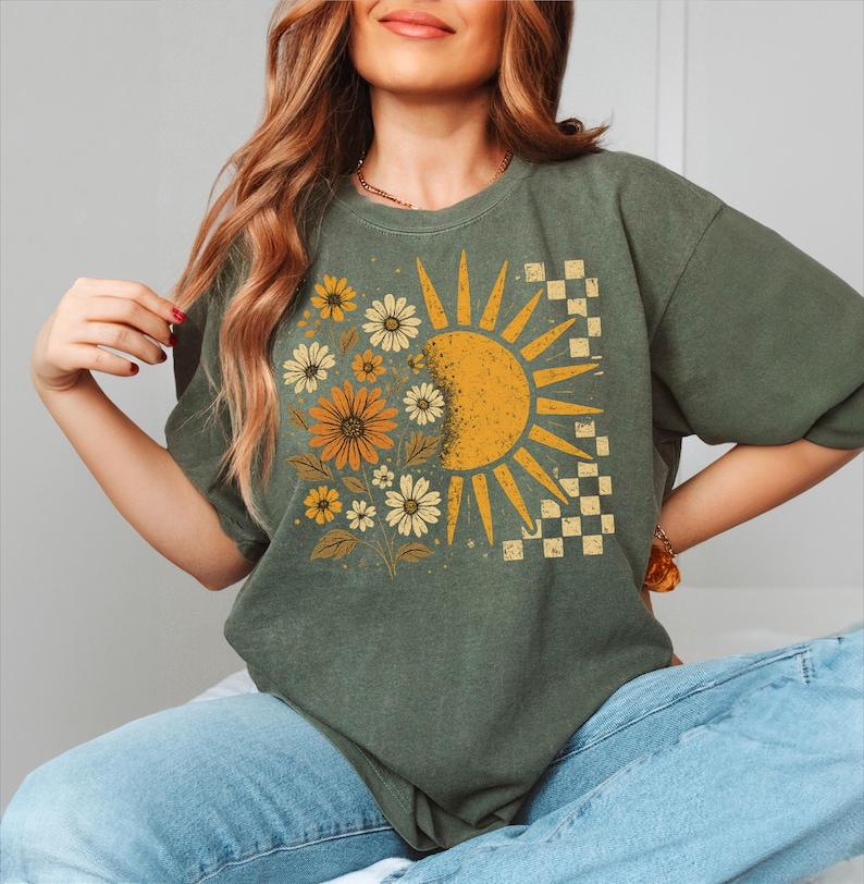 A woman wearing a green t-shirt with a sun and flower design, sitting with her hands on her hips.