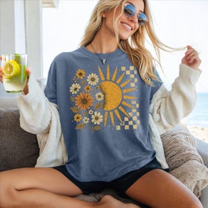 A woman wearing a blue t-shirt with a sun and flower design, sunglasses, and holding a drink is sitting on a couch.
