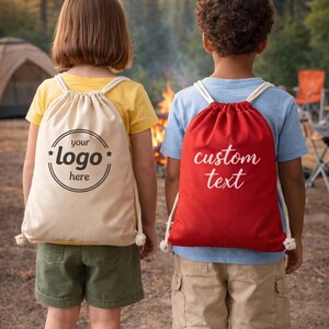Two children standing in front of a campfire, each carrying a bag with custom text printed on it.