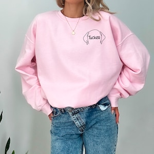 May include: A pink sweatshirt with a black outline of a dog's head and the name "Tucker" written underneath.
