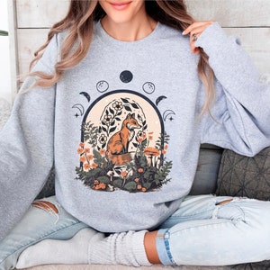 May include: A gray sweatshirt with a graphic print of a fox sitting in a forest with flowers and mushrooms. The graphic also includes a crescent moon and stars.