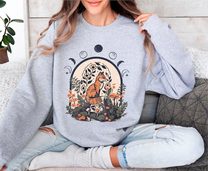 May include: A gray sweatshirt with a graphic print of a fox sitting in a forest with flowers and mushrooms. The graphic also includes a crescent moon and stars.