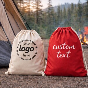 Two canvas bags, one white and one red, are placed on the ground near a tent and a campfire in a forested area.