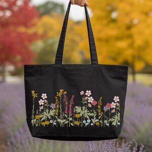 A hand holding a black tote bag with a floral pattern on it, set against a background of colorful autumn foliage.
