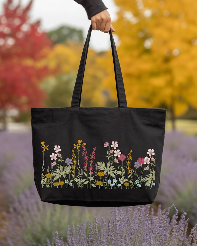 A hand holding a black tote bag with a floral pattern on it, set against a background of colorful autumn foliage.