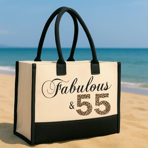 A beige tote bag with black straps and the text "Fabulous & 55" printed on it, placed on a sandy beach with the ocean in the background.
