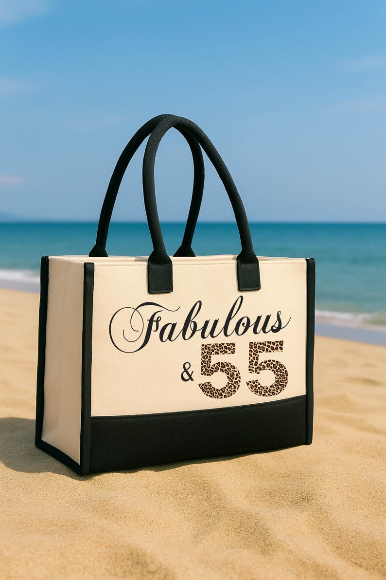 A beige tote bag with black straps and the text "Fabulous & 55" printed on it, placed on a sandy beach with the ocean in the background.