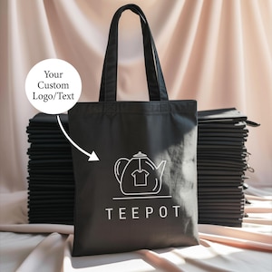 May include: Black tote bag with white graphic of a teapot and the word "TEE POT". The bag has long handles and is shown with a stack of similar bags. The image includes the text "Your Custom Logo/Text".
