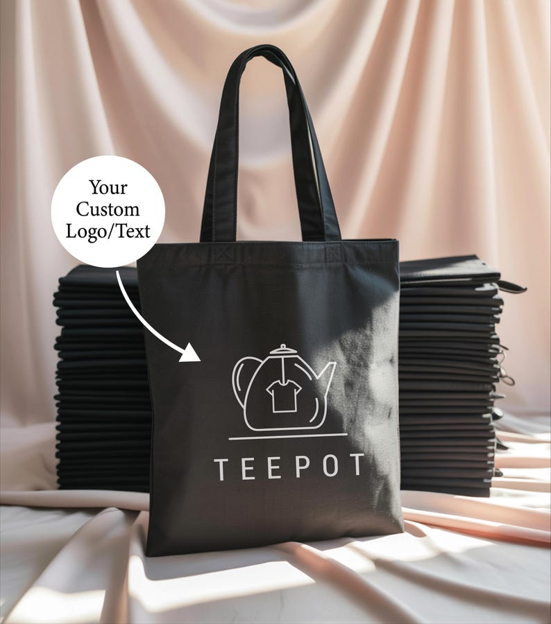 A black tote bag with a white teapot logo printed on it, placed on a surface with stacks of black fabric or cloth.
