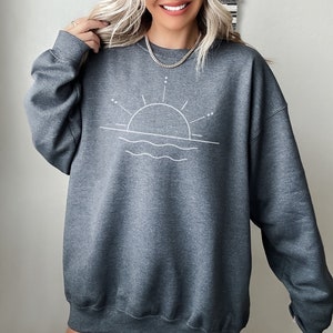 May include: A dark grey sweatshirt featuring a white graphic of a sun setting over water. The graphic is simple and minimalist, with a thin line drawing of the sun and waves.