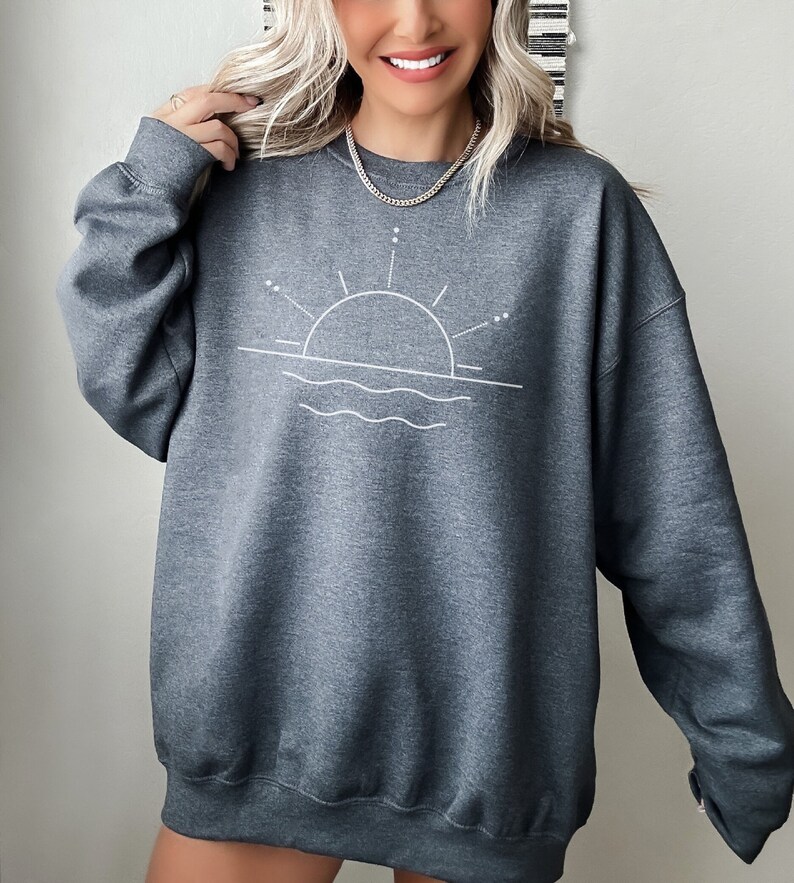 May include: A dark grey sweatshirt featuring a white graphic of a sun setting over water. The graphic is simple and minimalist, with a thin line drawing of the sun and waves.