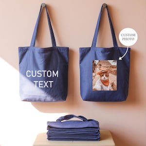 two blue tote bags hanging on a wall, one with a custom text printed on it and the other with a printed photo of a cat.