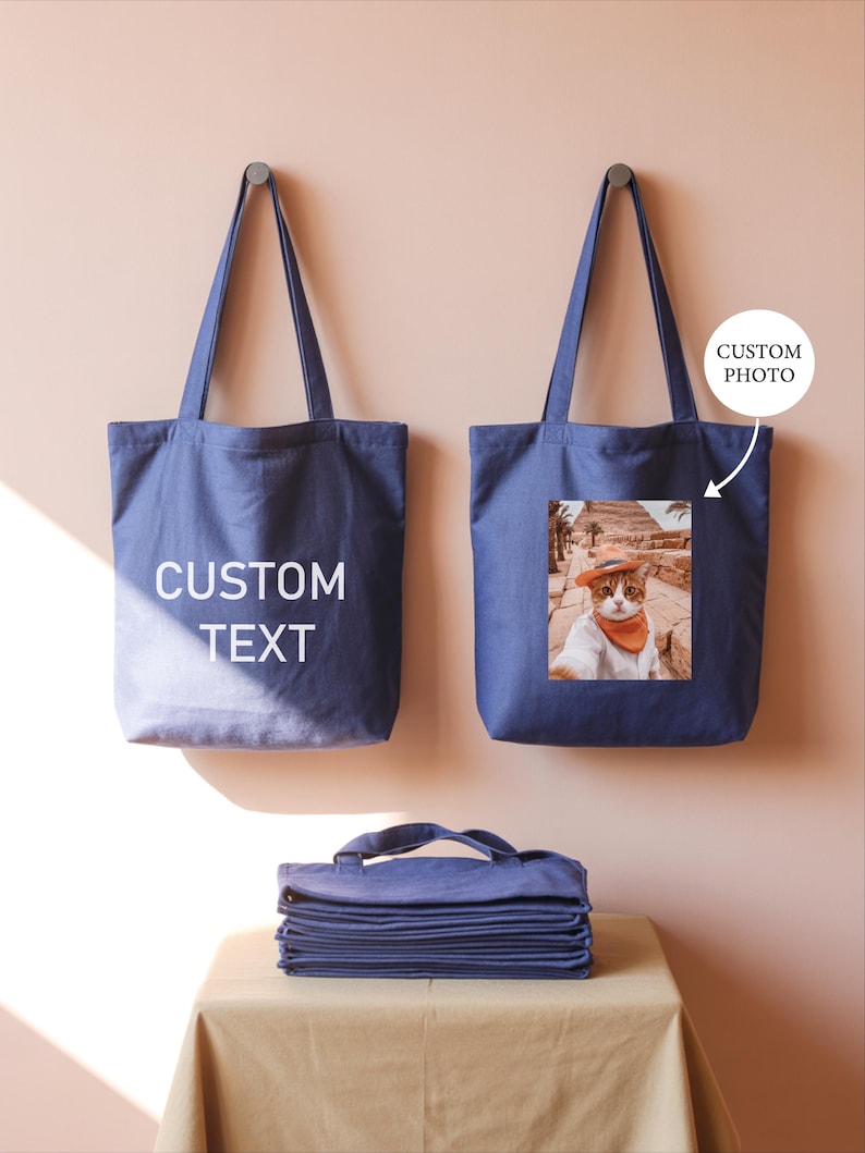 two blue tote bags hanging on a wall, one with a custom text printed on it and the other with a printed photo of a cat.