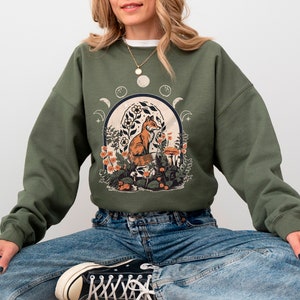 May include: A green sweatshirt with a graphic print of a fox sitting in a forest scene with flowers and mushrooms. The fox is orange and white. The scene is surrounded by a crescent moon and stars.