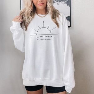 May include: White sweatshirt with a black line drawing of a sun setting over wavy water.